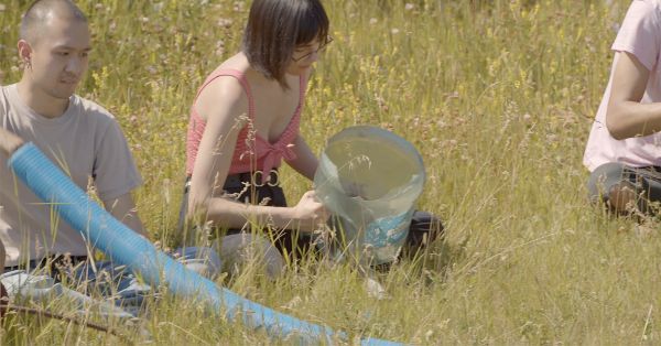 Medium shot of two people sitting on yellow grass. One holds a long blue plastic tube, the other holds the sawed-off bottom of a water cooler jug.