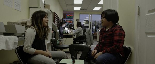 A Mohawk kid with long dark brown hair and highlights sits across from a Filipinx kid with short black hair and glasses in the back of a nail salon.