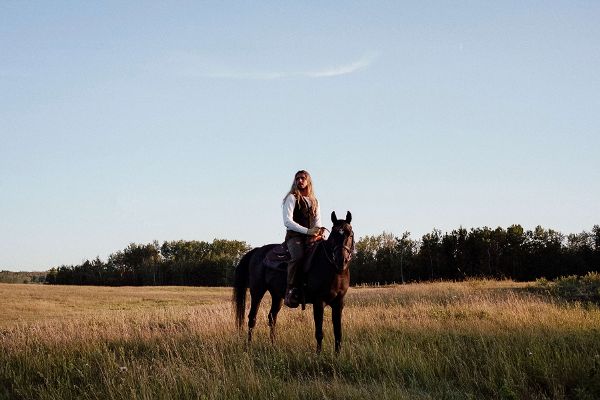 A Brown person with long blond hair wearing a white shirt and brown vest is on a horse, in a wide grassy area with trees in the distance.