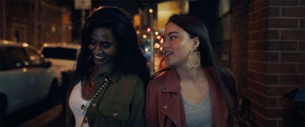 A dark-brown-skinned Tamil woman and lighter-brown-skinned Mohawk woman walk on a city street smiling and laughing together.