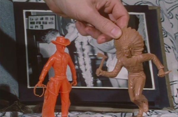 Image of a plastic orange cowboy figurine and brown “Indian” figurine in front of a black-and-white photo. A hand holds up the “Indian.”