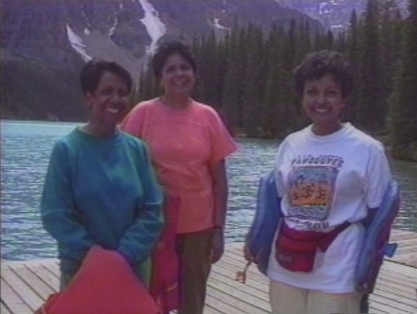 Three smiling middle-aged Brown women stand on a wooden pier at a lake with snow-peaked mountains in the background.