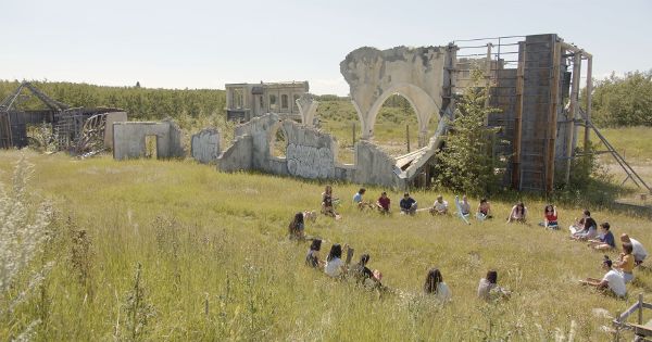 Long shot of a group of people sitting on long yellow grass on a clear sunny day, in front of concrete ruins.