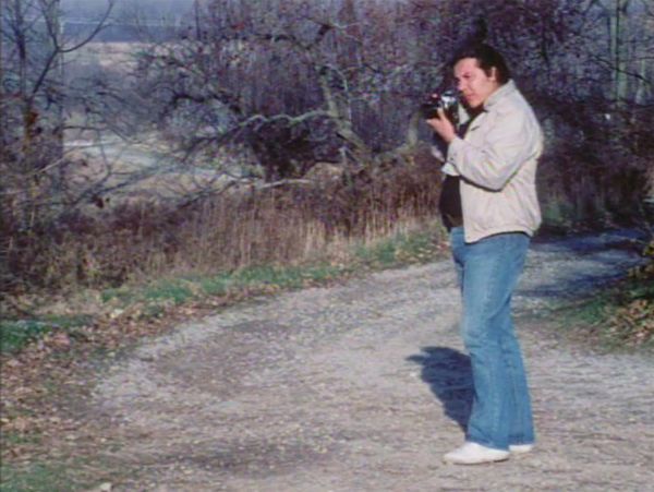 Thomas stands on the right taking a photo on a curved path amid fallen leaves and trees with bare branches.