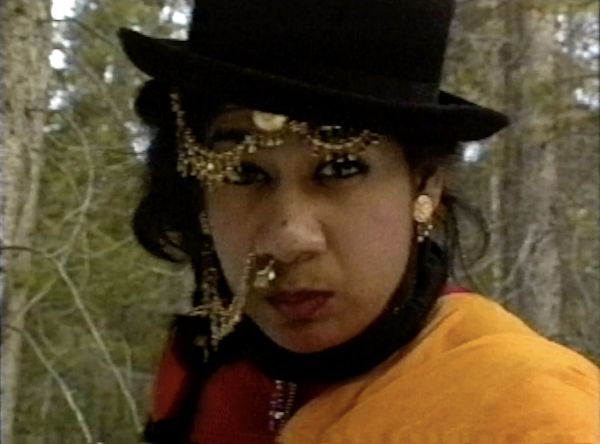 A Brown-skinned person looks straight into the camera. They have gold nose and forehead jewelry, a black bowler hat, and red and orange clothing.
