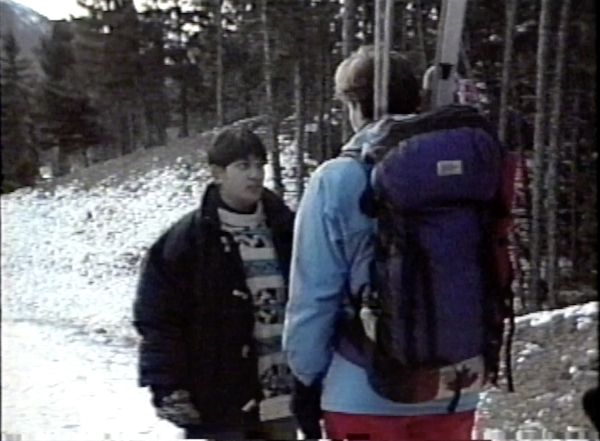On a snowy, forested, mountain path, a short-haired Brown person stands facing a taller, short-haired white person carrying ski gear in a backpack.