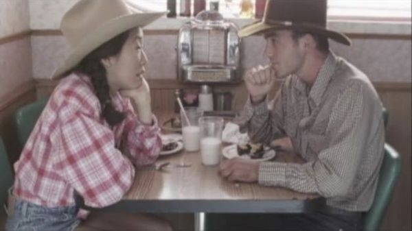 Oh has pigtails, a gingham shirt, and a cowboy hat. She sits across from a white man with a cowboy hat and checkered shirt.