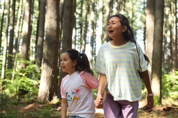 Two Mohawk children, one around 5–7 years old, the other preteen aged, stand crying out in a forest clearing as they look off into the distance.