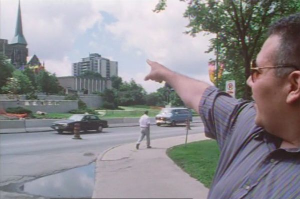 Close-up of Thomas with his face turned and pointing off in the distance. In the background are a church spire, high-rise building, and trees.