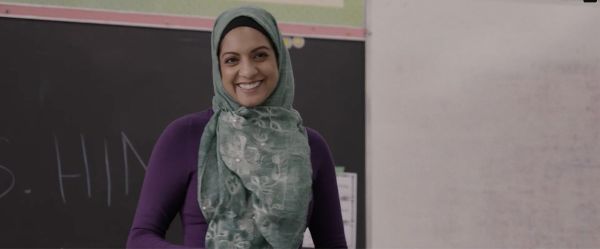A light-skinned Brown woman wearing a green hijab stands in front of a classroom blackboard smiling.