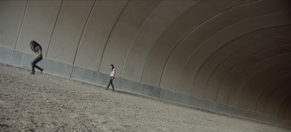 Slanted long shot of two individuals with long dark hair walking through a tunnel with gravel and greyish walls.
