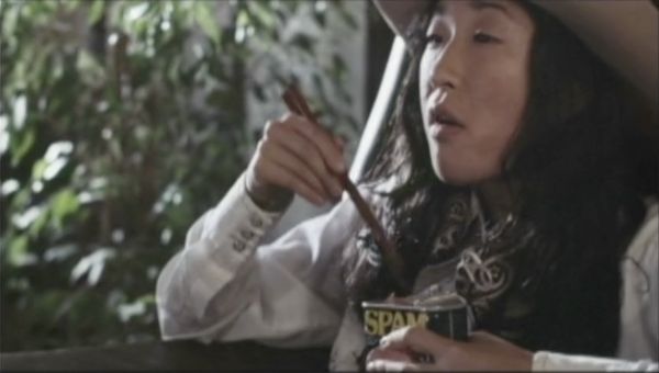 Close-up shot of actress Sandra Oh. She has long, dark, wavy hair, wears a cowboy hat, and is holding chopsticks and a can of Spam.