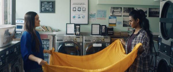A dark-brown-skinned Tamil woman and lighter-brown-skinned Mohawk woman get ready to fold a bright yellow piece of fabric at a laundromat.