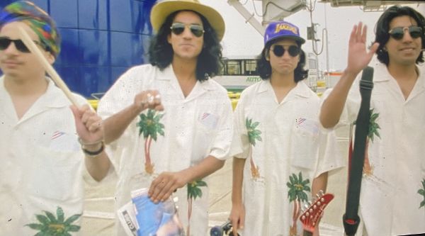 Four young Brown men wearing sunglasses and matching white shirts with palm trees stand outside at an airport.