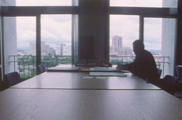 Backlit photo of Thomas sitting on a long table with large sheets of paper, in front of a window that looks onto buildings in the distance.