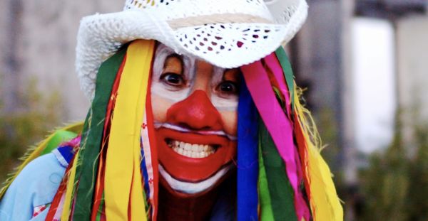 A person with a painted face grins. They are wearing a white crocheted cowboy hat and a wig with colored strips of fabric.
