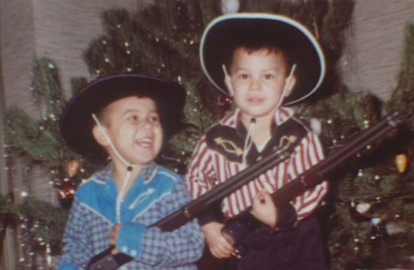 Childhood photo of Thomas dressed as a cowboy with his sibling. They are holding toy rifles and stand in front of a Christmas tree.