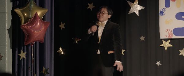 A chubby Filipinx kid with glasses, short hair, and a black tuxedo with gold bowtie sings into a microphone against a stage decorated with stars.